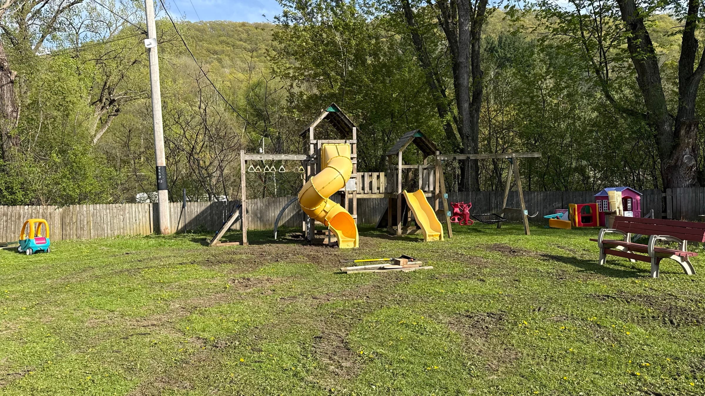 Children's playground at Camp Everyday Winona with a yellow tube slide, wooden climbing fort, swing set, and toddler playhouses on an open grassy lawn beneath the wooded bluffs